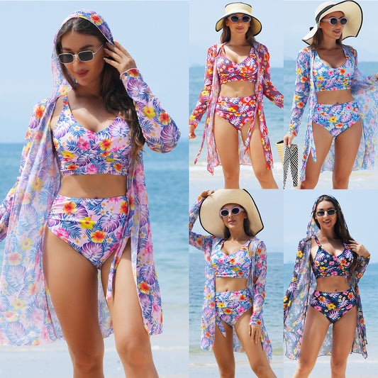 Woman in a red multicolor floral print three-piece swimsuit with high-waisted bikini and long-sleeve cover-up, straw hat, and sunglasses on a beach.