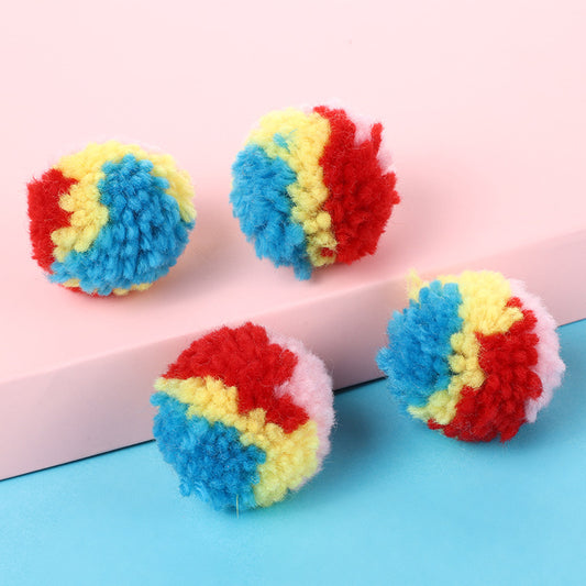 A hand holds a fluffy rainbow pom-pom ball toy, with another colorful ball in the background.