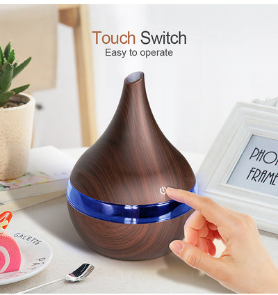 Dark wood grain LED essential oil diffuser with a blue light band and touch switch, shown with a hand operating it. Includes a bundle of 10 white filter sticks. - Image 7