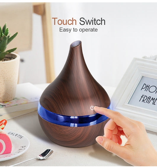 Dark wood grain LED essential oil diffuser with a blue light band and touch switch, shown with a hand operating it. Includes a bundle of 10 white filter sticks. - Image 7