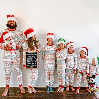 Family of four wearing matching green and white Christmas pajamas with fair isle print and red trim, sitting on a bed.