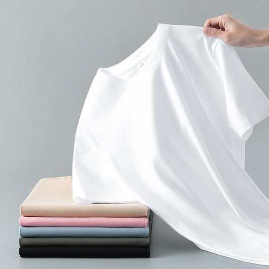 Light gray short-sleeve t-shirt with a round neck, held up to show its smooth fabric. A stack of folded t-shirts in various colors is visible in the background. - Image 3