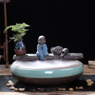Ceramic flowing water ornament with a meditating monk figurine, small plant, and misting humidifier in a teal and brown bowl. - Image 6
