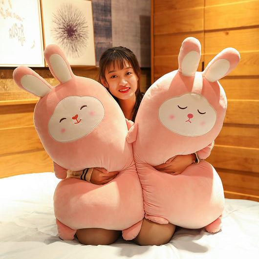 Pink rabbit plush doll with closed eyes, soft and elongated, standing on a bed.