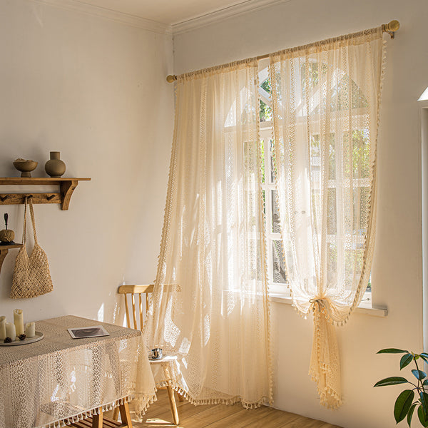 Light beige sheer lace curtains with bohemian patterns and tassel trim, hung on a wooden rod in a sunny room.