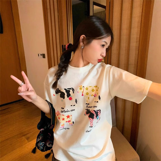 White loose-fit short-sleeve top with colorful retro cartoon cow print, modeled by a woman with plaid pants.