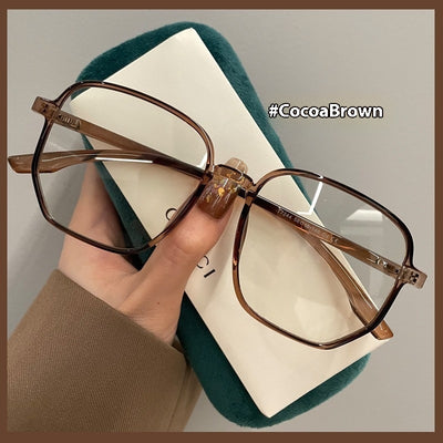 Mocha brown irregular frame glasses with clear lenses, held over a dark green velvet glasses case. - Image 5