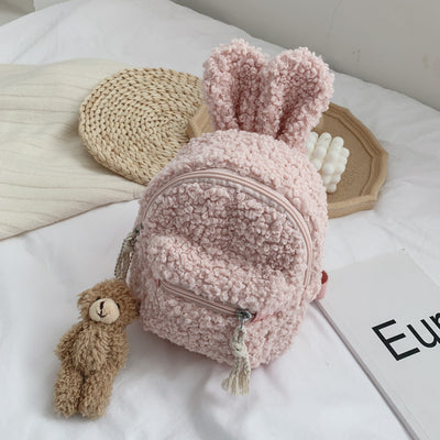 Pink fuzzy mini backpack with bear ears and a small white teddy bear keychain on a white bed. - Image 4