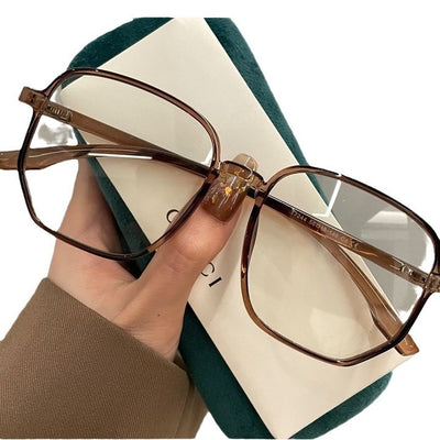 Mocha brown irregular frame glasses with clear lenses, held over a dark green velvet glasses case. - Image 2