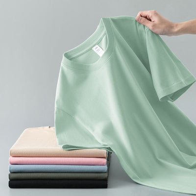 Light gray short-sleeve t-shirt with a round neck, held up to show its smooth fabric. A stack of folded t-shirts in various colors is visible in the background. - Image 13