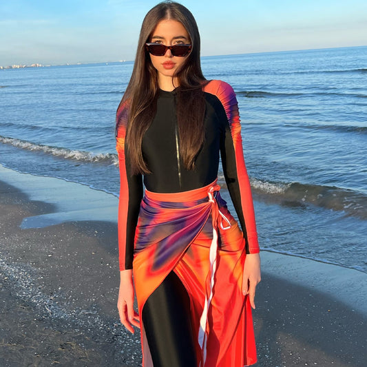 Woman in a black long-sleeve rash guard, black swim leggings, and a vibrant tie-dye sarong on a sunny beach.