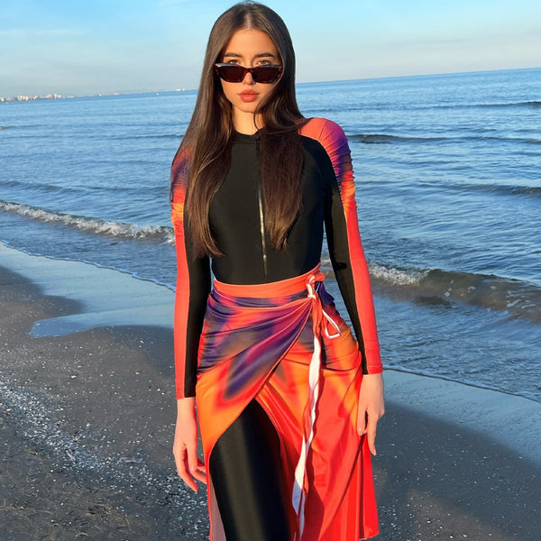 Woman in a black long-sleeve rash guard, black swim leggings, and a vibrant tie-dye sarong on a sunny beach.