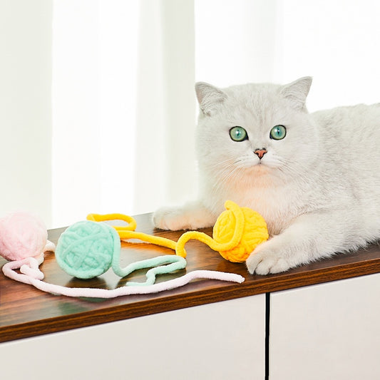 Colorful plush yarn ball cat toy in pink, blue, and white, with a long trailing strand, on a light background.