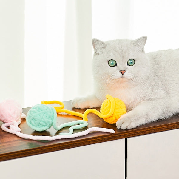 Colorful plush yarn ball cat toy in pink, blue, and white, with a long trailing strand, on a light background.