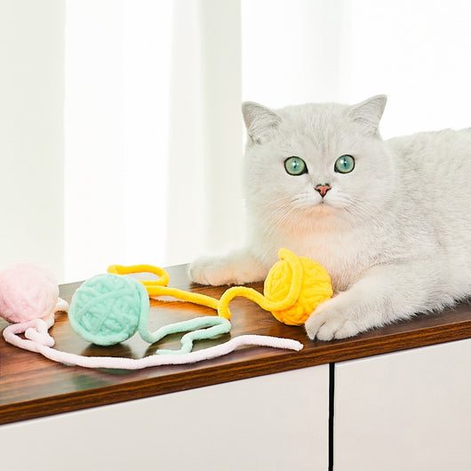 Colorful plush yarn ball cat toy in pink, blue, and white, with a long trailing strand, on a light background.