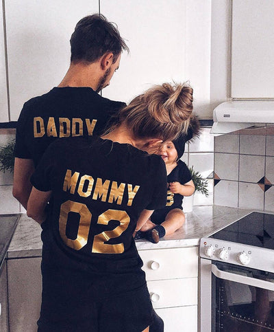 Child's black short-sleeved t-shirt with 'BABY 04' in gold print on the back, worn by a toddler in grass. - Image 3