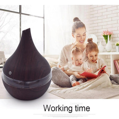 Dark wood grain LED essential oil diffuser with a blue light band and touch switch, shown with a hand operating it. Includes a bundle of 10 white filter sticks. - Image 6
