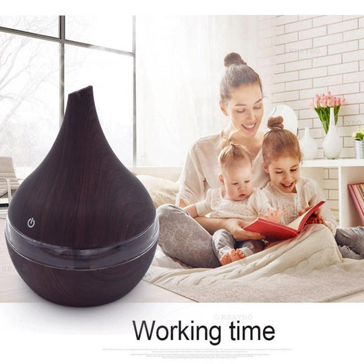 Dark wood grain LED essential oil diffuser with a blue light band and touch switch, shown with a hand operating it. Includes a bundle of 10 white filter sticks. - Image 6