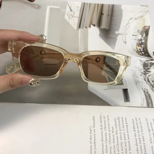 Square retro tortoise shell sunglasses with dark lenses, held by a hand against a blurred magazine background.