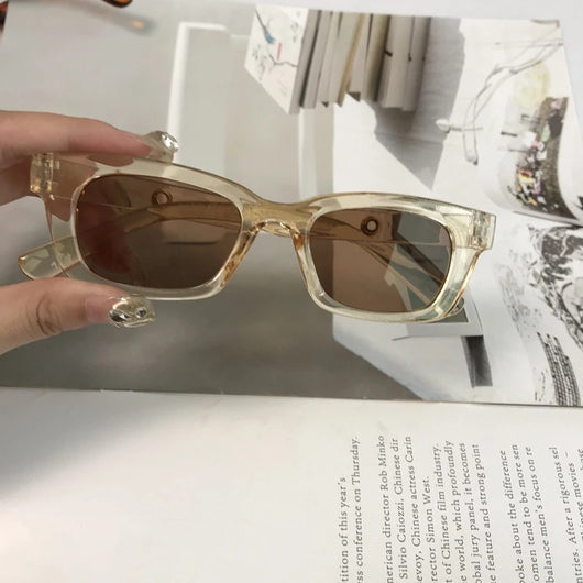 Square retro tortoise shell sunglasses with dark lenses, held by a hand against a blurred magazine background.
