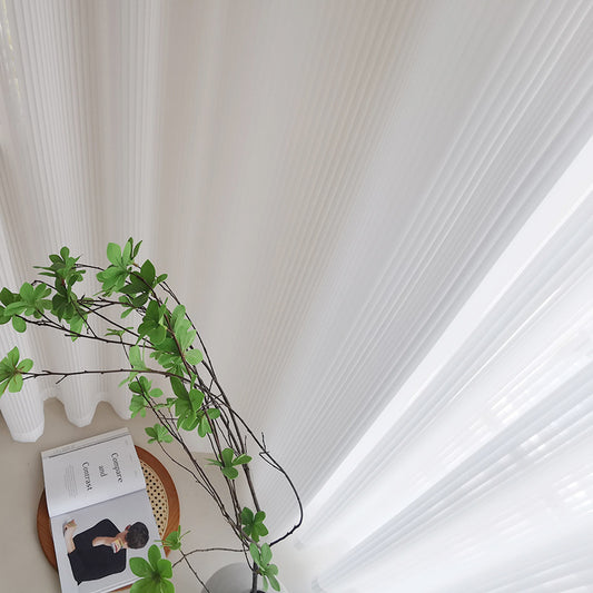 White vertical blinds with a pleated texture, allowing sunlight to filter through and cast shadows on an open magazine. - Image 2