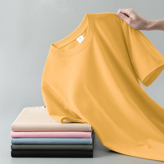 Light gray short-sleeve t-shirt with a round neck, held up to show its smooth fabric. A stack of folded t-shirts in various colors is visible in the background. - Image 12