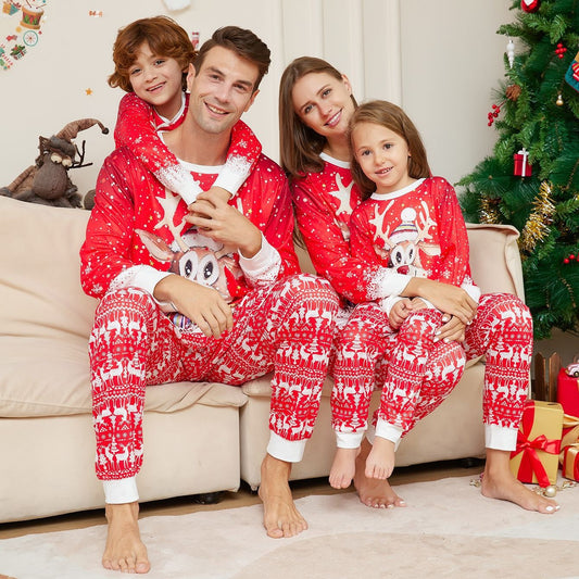 Family of four wearing matching red Christmas pajamas with reindeer and snowflake patterns, sitting on a sofa.