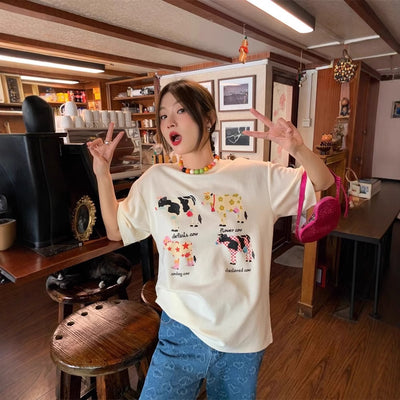 White loose-fit short-sleeve top with colorful retro cartoon cow print, modeled by a woman with plaid pants. - Image 4
