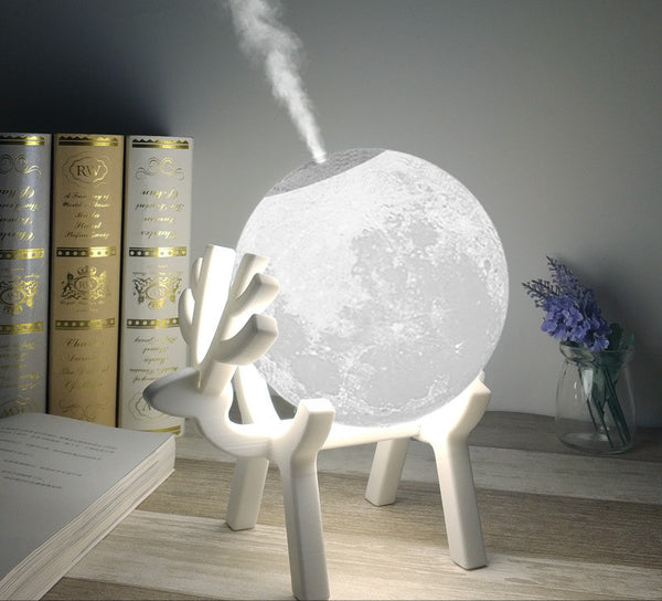 Moon-shaped humidifier and night light with a white deer stand, emitting mist, on a wooden table with books.