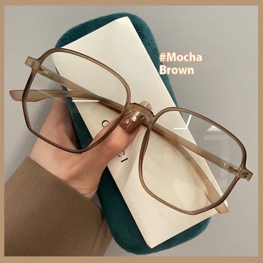 Mocha brown irregular frame glasses with clear lenses, held over a dark green velvet glasses case. - Image 8