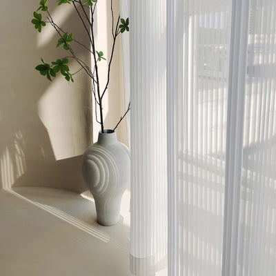 White vertical blinds with a pleated texture, allowing sunlight to filter through and cast shadows on an open magazine.