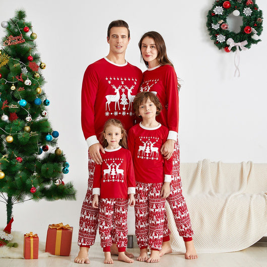 Family of four wearing matching red Christmas pajamas with white reindeer and tree patterns, standing by a decorated tree.