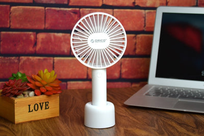 White ORICO FH1 handheld USB fan with charging dock, USB cable, and user manuals on a wooden desk.