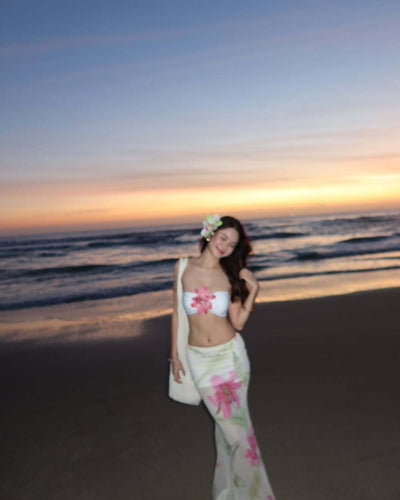 Woman in a white tube top with pink flower and a sheer floral maxi skirt, posing on a beach at sunset. - Image 15