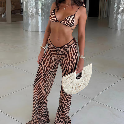 Woman in a brown and black animal print bikini with matching sheer wide-leg cover-up pants, holding a woven straw handbag. - Image 4