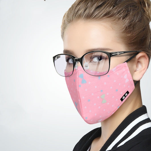 Woman wearing a pink cotton face mask with light blue polka dots and bow patterns, paired with black-framed glasses. - Image 8