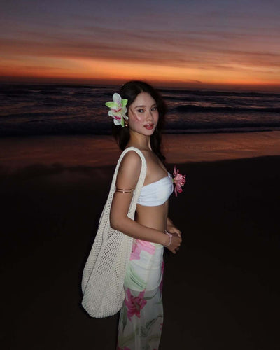 Woman in a white tube top with pink flower and a sheer floral maxi skirt, posing on a beach at sunset. - Image 13