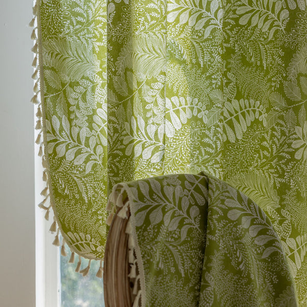 Green curtain with white leafy botanical pattern and cream tassels, hanging in a window with natural light.