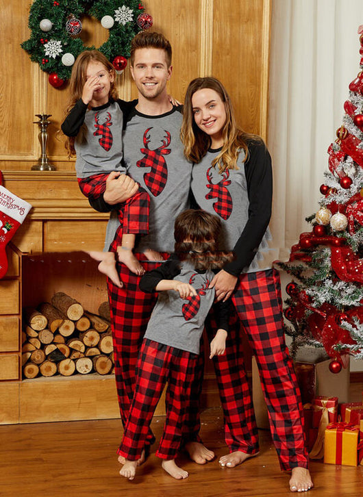 Family of four wearing matching gray and red plaid Christmas pajamas with reindeer graphic, standing by a festive fireplace and Christmas tree.