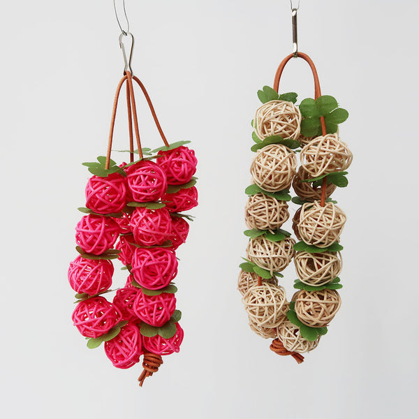 Pink woven rattan ball parrot chew toy with green leaf accents, hanging from a metal hook.