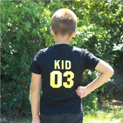 Child's black short-sleeved t-shirt with 'BABY 04' in gold print on the back, worn by a toddler in grass. - Image 7