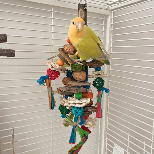 Hanging parrot toy made from natural wood, corn husk, and nuts with colorful strings and beads. A small yellow bird perches on the toy.