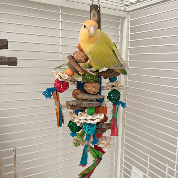 Hanging parrot toy made from natural wood, corn husk, and nuts with colorful strings and beads. A small yellow bird perches on the toy.
