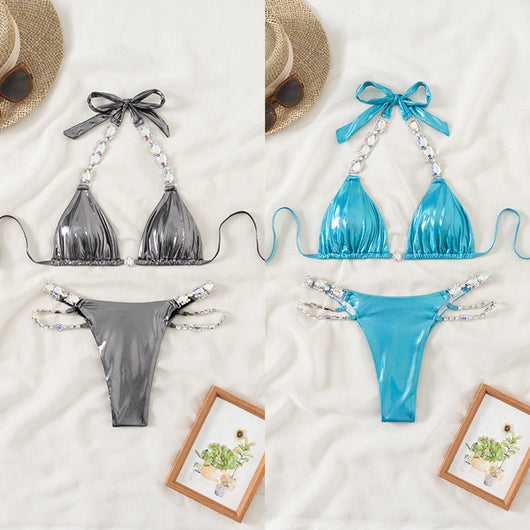 Shiny blue two-piece bikini with rhinestone-embellished halter top and high-cut bottom, laid flat with a straw hat and sunglasses.