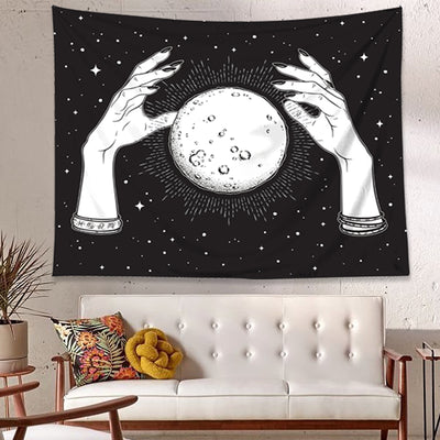 Black and white tapestry featuring two hands holding a full moon against a starry night sky, hanging on a white wall.