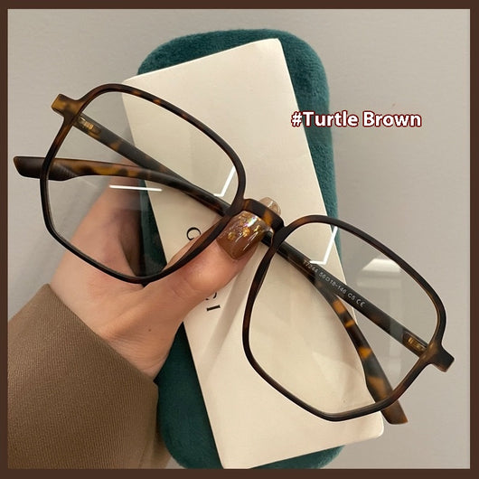 Mocha brown irregular frame glasses with clear lenses, held over a dark green velvet glasses case. - Image 3