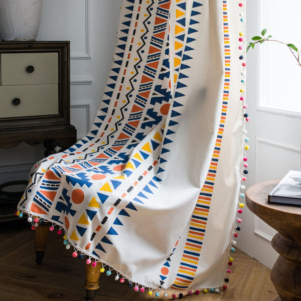 Bohemian cotton and linen curtain with colorful geometric prints and fur ball trim, hanging in a bright room.