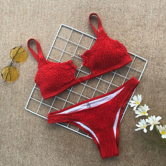 Red pleated cutout bikini set with a triangle top and thong bottom, displayed with yellow sunglasses and white flowers.
