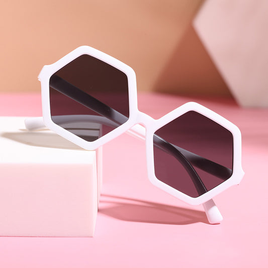 Red hexagonal children's sunglasses with dark lenses on a pink surface with a white block.