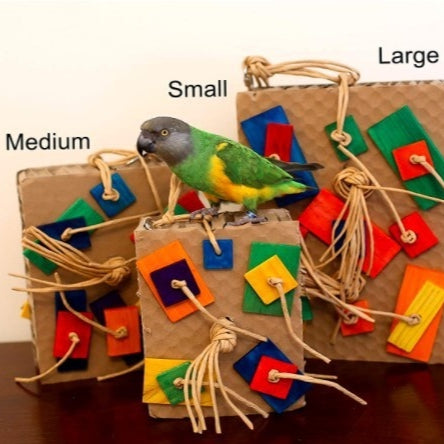 A green and yellow parrot on a wooden perch interacts with a colorful cardboard and wood block nibbling toy.